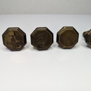 May include: Four octagonal cabinet knobs with a weathered brass finish. Each knob features a flat, multi-faceted top surface with beveled edges and a dark, aged metallic patina, arranged in a horizontal row against a plain white background.