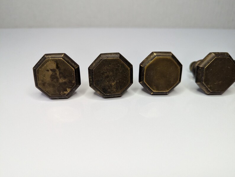 May include: Four octagonal cabinet knobs with a weathered brass finish. Each knob features a flat, multi-faceted top surface with beveled edges and a dark, aged metallic patina, arranged in a horizontal row against a plain white background.
