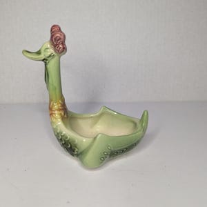 以下が含まれることがあります： スタイリッシュなアヒルの形をしたヴィンテージセラミックプランター。プランターは主に薄緑色で、内側はクリーム色です。アヒルは長い首、赤い鶏冠、装飾的なディテールが特徴です。