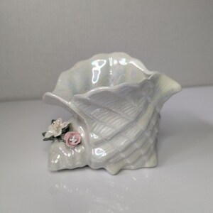 May include: A ceramic shell-shaped vessel with an iridescent, pearlescent finish. The exterior features a textured, ribbed surface and is decorated with small, delicate sculpted flowers in shades of white and soft pink on the lower side.