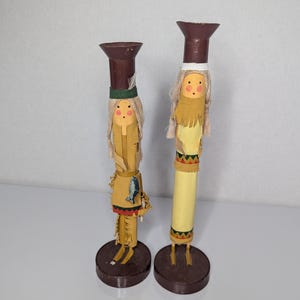May include: Two tall, cylindrical decorative figurines standing on circular brown bases. Each figure features a painted face, light-colored hair, and a tan garment with fringe details. The figures are topped with brown, flared, cup-like structures.