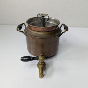 May include: A vintage copper vessel featuring two side handles with black wooden grips, a brass spigot with a black wooden handle, and an open top revealing a central metal heating element or burner mechanism inside the main cylindrical body.
