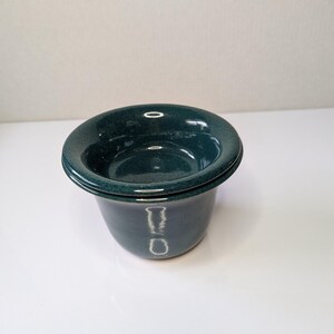 May include: A dark teal ceramic butter keeper with a glossy finish. The vessel consists of a deep base and a separate lid with a wide, flared rim. The glaze has a subtle speckled texture throughout, reflecting light on its smooth, curved surface.