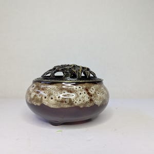 May include: A ceramic incense burner with a dark brown base and a speckled, light brown and white upper section. The lid is dark metal with a decorative, openwork design. The burner has three small feet.