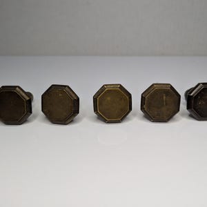 May include: A set of six brass-toned metal cabinet knobs, each featuring an octagonal face with a flat surface and beveled edges. The knobs are arranged in a horizontal row, displaying a weathered, metallic finish with subtle variations in tone.