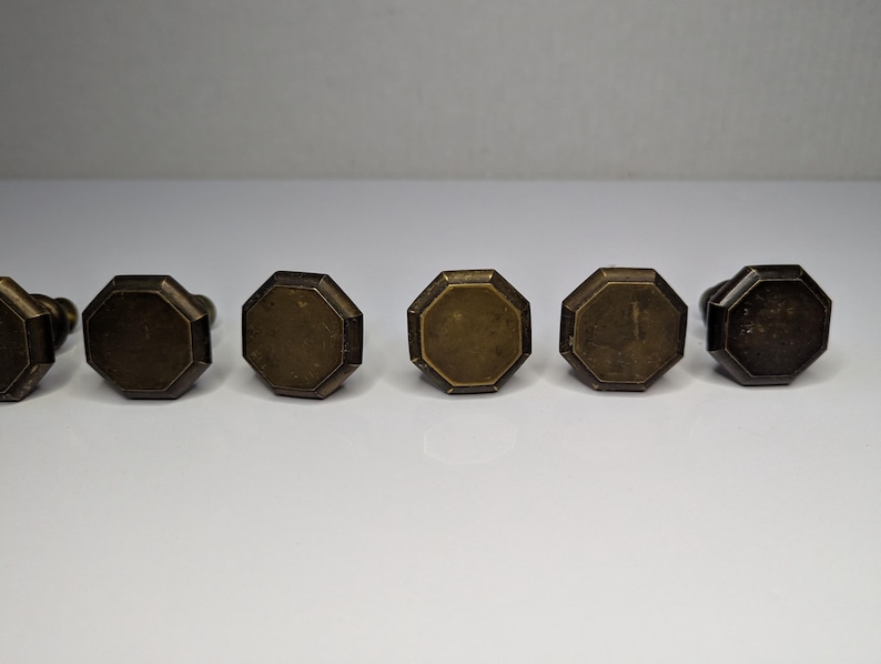May include: A set of six brass-toned metal cabinet knobs, each featuring an octagonal face with a flat surface and beveled edges. The knobs are arranged in a horizontal row, displaying a weathered, metallic finish with subtle variations in tone.
