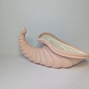 May include: A light pink, horn-shaped planter with a textured exterior. The interior is white. The planter has a curved, spiral design and a wide opening. This decorative piece is suitable for displaying plants or flowers.