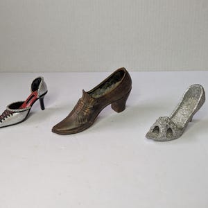 Puede incluir: Tres zapatos decorativos en miniatura. Un tacón alto plateado y negro con interior rojo, un zapato con tacón marrón y un tacón alto plateado con purpurina y punta abierta. Los zapatos están colocados sobre una superficie blanca.