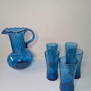 Könnte beinhalten: Eine blaue Glaskaraffe mit gerüschtem Rand und Griff, zusammen mit fünf passenden hohen Gläsern. Das Glaswaren hat ein Wirbeldesign, das ein strukturiertes Aussehen erzeugt. Das Set wird auf einer weißen Oberfläche präsentiert.