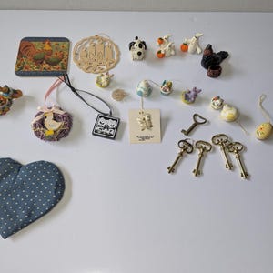 Könnte beinhalten: Eine Sammlung kleiner Dekorationsartikel, darunter ein herzförmiger Stoffbeutel in Blau, ein Untersetzer mit Hahnmotiv, ein hölzerner Kerzenschmuck, verschiedene Tierfiguren, ein schwarz-weißer quadratischer Anhänger und vier antike Metallschlüssel.