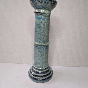 May include: A tall, ceramic pillar candle holder with a textured, speckled teal glaze. The base and top have dark blue accents. The pillar is cylindrical with a flared top, and the base has concentric rings.
