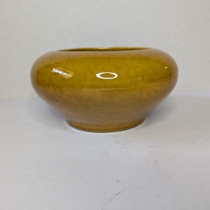 May include: A small, round ceramic bowl with a glossy mustard yellow glaze. The bowl features a smooth, curved silhouette with a wide opening and a slightly tapered base, showcasing a speckled texture throughout the surface of the pottery.