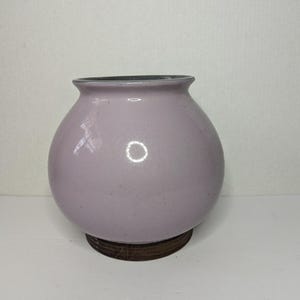May include: A round, lavender-colored ceramic vase with a dark rim. The vase sits on a dark brown wooden base. The vase has a smooth, glossy finish.