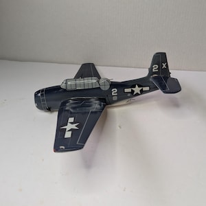 May include: A dark blue toy airplane with white star and cross markings on the wings and tail. The plane has the number "2" on the fuselage and tail, and the letter "X" on the tail. The plane is a vintage model.