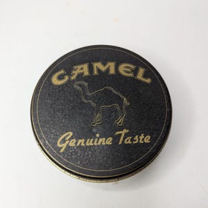 May include: A round, black tin with gold lettering and a camel silhouette. The word "CAMEL" arches across the top, with "Genuine Taste" below. The tin has a textured surface and a gold rim.