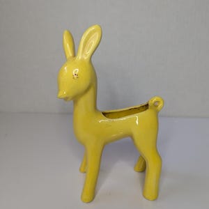 May include: A bright yellow ceramic deer-shaped planter with a glossy finish. The deer is standing in profile with upright ears and a small curled tail. The opening for the planter is located on the back of the deer, featuring a smooth rim.