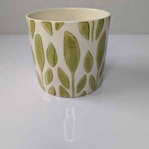 May include: A cylindrical ceramic planter featuring an off-white base color with a repeating pattern of sage green leaves. The leaves are arranged in a vertical orientation around the exterior surface of the pot, creating a consistent botanical design.