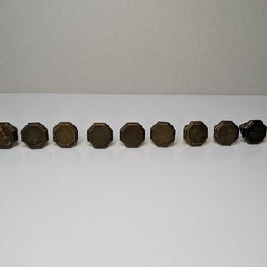 May include: A set of nine octagonal cabinet knobs made of aged brass with a metallic finish. Each knob features a flat, multi-sided face and a beveled edge, arranged in a horizontal line against a plain, neutral background for clear visibility.