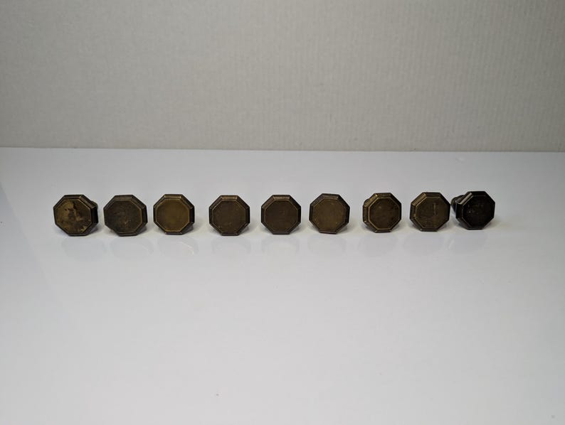 May include: A set of nine octagonal cabinet knobs made of aged brass with a metallic finish. Each knob features a flat, multi-sided face and a beveled edge, arranged in a horizontal line against a plain, neutral background for clear visibility.