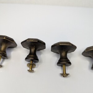 May include: A set of four antique brass-toned metal cabinet knobs, each featuring an octagonal top surface and a tapered base. Each knob includes a threaded metal screw and a small circular washer attached to the underside for installation.