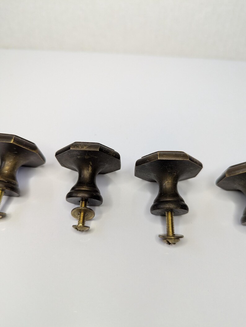 May include: A set of four antique brass-toned metal cabinet knobs, each featuring an octagonal top surface and a tapered base. Each knob includes a threaded metal screw and a small circular washer attached to the underside for installation.