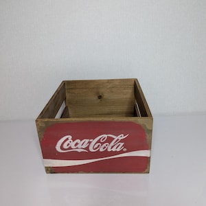 Puede incluir: Una caja de madera rectangular con un acabado marrón natural. El panel frontal presenta un gráfico rectangular rojo con el logotipo blanco de Coca-Cola y una línea curva blanca debajo. La caja tiene asas laterales abiertas y un interior hueco.