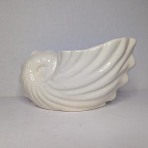 May include: A white ceramic seashell-shaped planter. The planter has a spiral design on one end and a wavy, open top. The smooth, glossy surface reflects light, highlighting the curves and details of the nautical-themed decor.