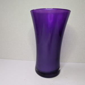 May include: A tall, elegant vase in a deep purple hue. The vase has a flared top and a gently curved body, creating a graceful silhouette. The glass appears smooth and reflective, with a dark base.