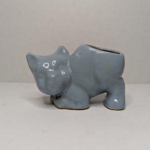 Può includere: Una fioriera a forma di gatto in ceramica azzurra. La fioriera ha una parte superiore scavata per la piantumazione ed è progettata in una forma di gatto stilizzata. Il gatto è in posizione accovacciata, con la testa in su e la coda in giù. La ceramica ha una finitura lucida.