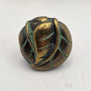 May include: A round, bronze-colored cabinet knob with a leaf design. The knob has a gold finish with green accents in the leaf veins. The hardware is likely made of metal and is suitable for furniture or decorative projects.