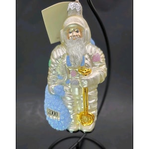 May include: A pearlescent Christmas ornament depicting Santa Claus in a spacesuit, holding a golden shovel and a blue sack with "2000" on it. The ornament has a white beard and is accented with pink and blue details. It is suspended from a black stand.