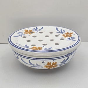 May include: White ceramic soap dish with a floral design. The dish features a round shape with a perforated top and a bowl-like base. The design includes orange flowers and blue leaves, with blue trim.