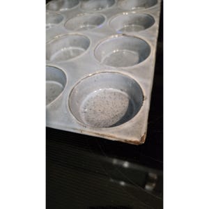 Puede incluir: Un molde para muffins de cerámica con doce moldes circulares. La bandeja es de color gris claro con detalles moteados en el interior de cada molde. Los bordes tienen un acabado ligeramente más oscuro y rústico.