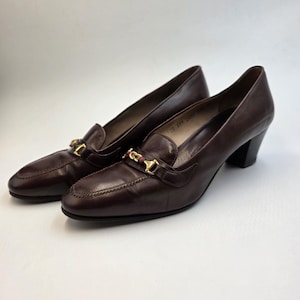 May include: A pair of dark brown leather heeled loafers. Each shoe features a gold-colored metal accent with a red and gold stripe. The heels are a solid black color.