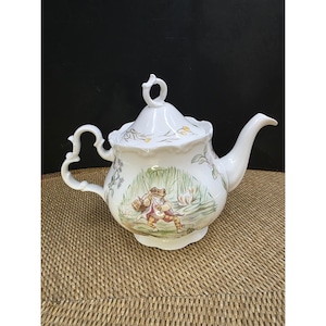 May include: White ceramic teapot with a lid and curved spout. The teapot features a whimsical illustration of a frog in a pond scene. The handle is uniquely shaped. Floral and plant details are painted on the teapot.