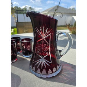 May include: A ruby red crystal jug with a clear handle and matching glasses. The jug and glasses feature a starburst and geometric design. The set is displayed on a light-coloured surface.