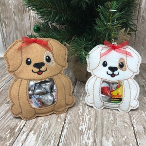 May include: Two felt dog ornaments with clear plastic windows for holding treats. The brown dog has a red bow and the white dog has a red bow. Both dogs have brown eyes and noses.