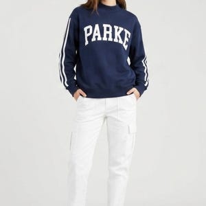 以下が含まれることがあります： 白いブロック文字で「PARKE」と書かれたネイビーブルーのスウェットシャツ。スウェットシャツの袖には白いストライプが入っています。モデルは白いカーゴパンツと白いスニーカーを着用しています。