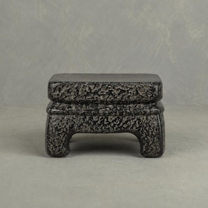 May include: A small, dark gray decorative stand with a textured surface. The stand has a rectangular top and a detailed base with four short legs. The stand is made of a stone-like material and is suitable for displaying small objects.