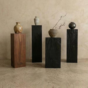 May include: Four rectangular wooden pedestals of varying heights and finishes, including one natural brown wood and three charred black wood columns. Each pedestal supports a unique ceramic vessel, including a tan vase, a grey vase, and a black sphere.