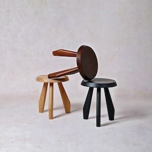 May include: Three wooden stools with round seats and three angled legs. One stool is light natural wood, one is dark brown wood, and one is painted black. The dark brown stool is positioned at an angle, resting on the other two stools.