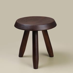 May include: A dark brown wooden stool with a round seat and three legs. The stool has a simple, minimalist design and is made of solid wood. The wood grain is visible on the seat and legs.