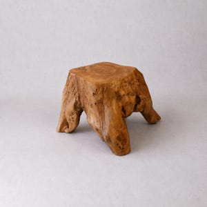 May include: A natural wood stump with a square top and three sturdy legs. The light brown wood has a textured surface with visible knots and grain, creating a rustic and organic aesthetic. This unique piece could be used as a small table or decorative accent.