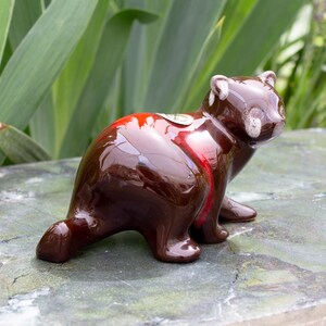 May include: A brown and red glazed ceramic figurine of a raccoon. The figurine has white accents on the face and a glossy finish.