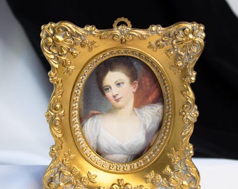 May include: A gold-colored, ornate picture frame with an oval portrait of a woman in a white dress. The frame has detailed floral and scrollwork designs. The portrait features a woman with light skin, brown hair, and blue eyes.