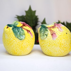 May include: A pair of ceramic lemon-shaped salt and pepper shakers. The shakers are yellow with a textured surface and feature green leaves and pink accents. The shakers are designed to resemble lemons.