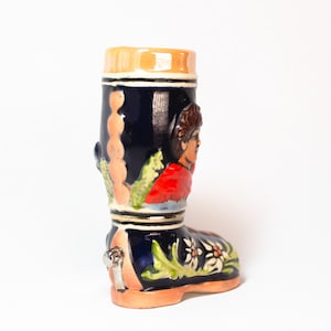 May include: A ceramic boot-shaped beer stein with a blue and white design. The stein features a painted scene of a person in a red and blue outfit.