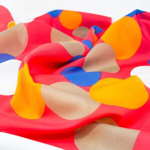 May include: A vibrant red fabric with a pattern of large, irregularly shaped circles in orange, yellow, blue, white, and tan. The fabric is soft and flowing, with visible folds and creases, suggesting a scarf or similar textile.