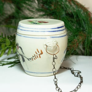 May include: A white ceramic jar with a blue and white floral design and a metal chain attached to the lid. The jar has a small bird painted on the side.