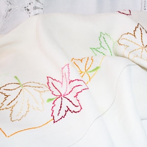 May include: White linen tablecloth with embroidered autumn leaves in shades of green, orange, pink, and red.